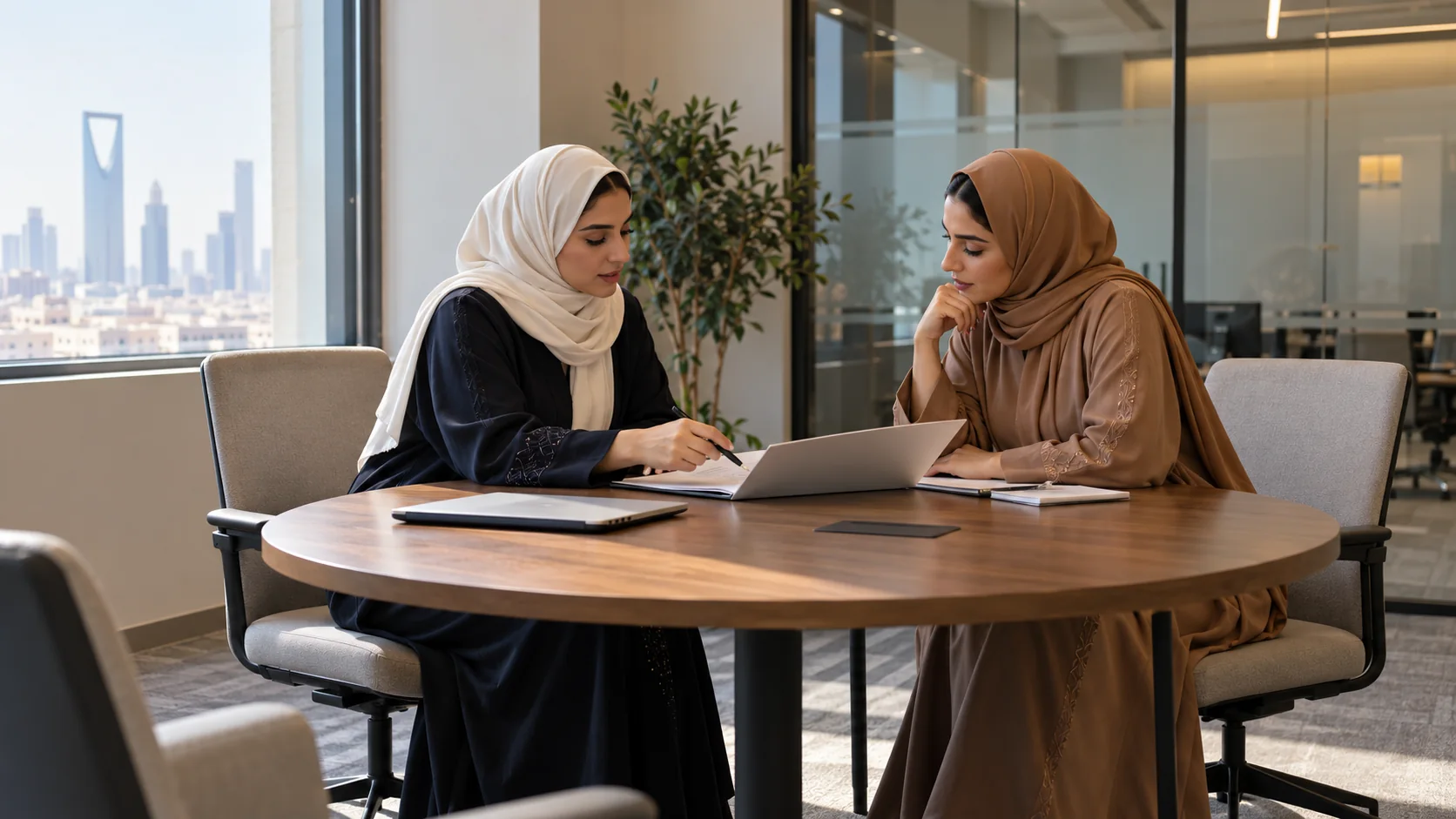 Two women in navy and caramel abayas discussing an open planning folder at a round office meeting table